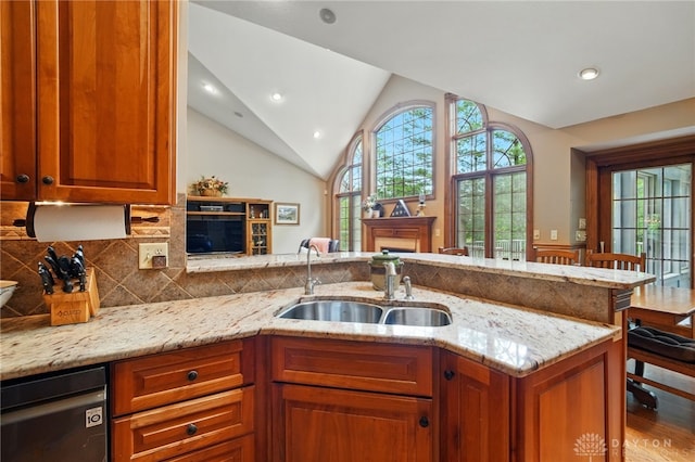 kitchen featuring lofted ceiling, dishwashing machine, brown cabinets, light stone counters, and a sink