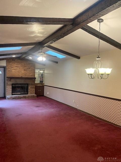 unfurnished living room with ceiling fan with notable chandelier, a wainscoted wall, a fireplace, lofted ceiling with beams, and dark colored carpet