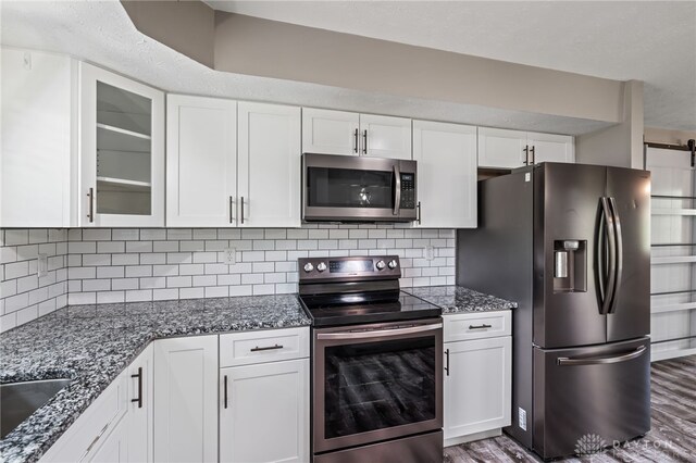 kitchen featuring stainless steel appliances, white cabinetry, dark stone counters, glass insert cabinets, and wood finished floors