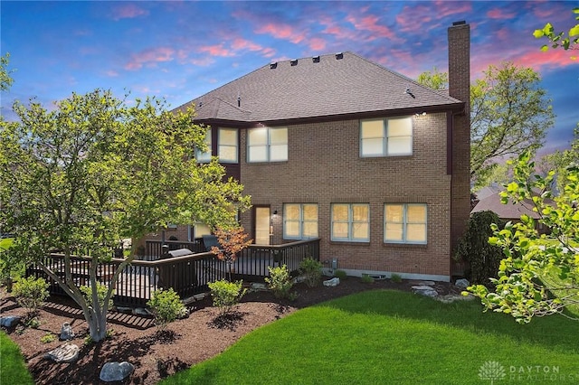 rear view of house featuring a chimney, a lawn, a wooden deck, brick siding, and roof with shingles