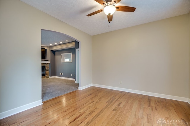 empty room with arched walkways, ceiling fan, baseboards, light wood-type flooring, and a stone fireplace