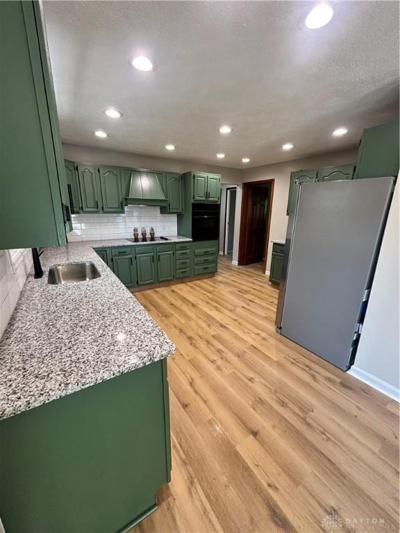 kitchen with green cabinetry, custom range hood, a sink, light wood-style floors, and decorative backsplash