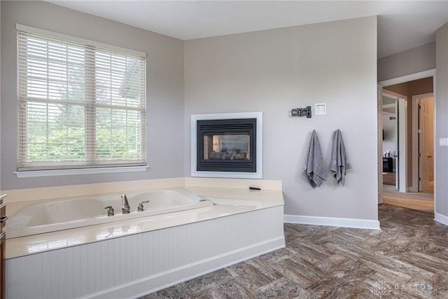 bathroom featuring a bath, plenty of natural light, a glass covered fireplace, and baseboards