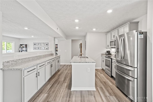 kitchen with stainless steel appliances, a sink, a kitchen island with sink, a textured ceiling, and light wood finished floors