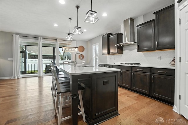 539 Bellasera Dr, Sugarcreek Twp, OH 45440 - kitchen featuring wall chimney range hood, decorative backsplash, light countertops, a center island with sink, and light wood finished floors