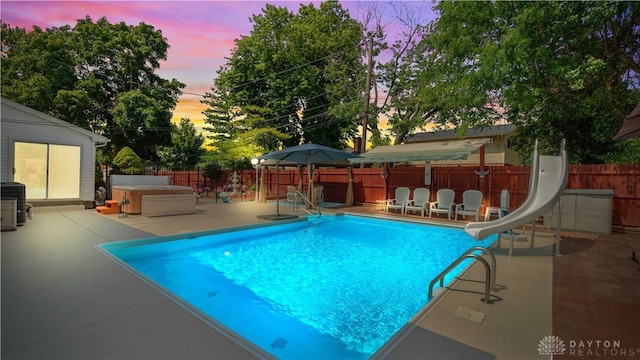 429 Rader Dr, Vandalia, OH 45377 - pool at dusk featuring a water slide, a patio area, a fenced backyard, and a covered hot tub
