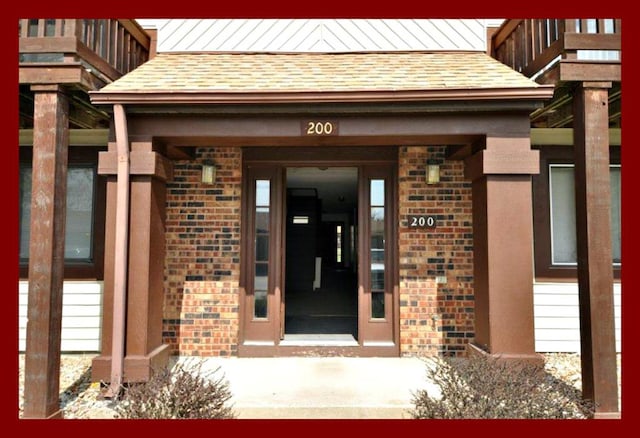 view of exterior entry with a shingled roof and brick siding