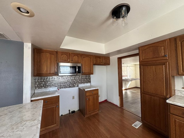 kitchen with a tray ceiling, stainless steel microwave, decorative backsplash, dark wood-type flooring, and brown cabinetry