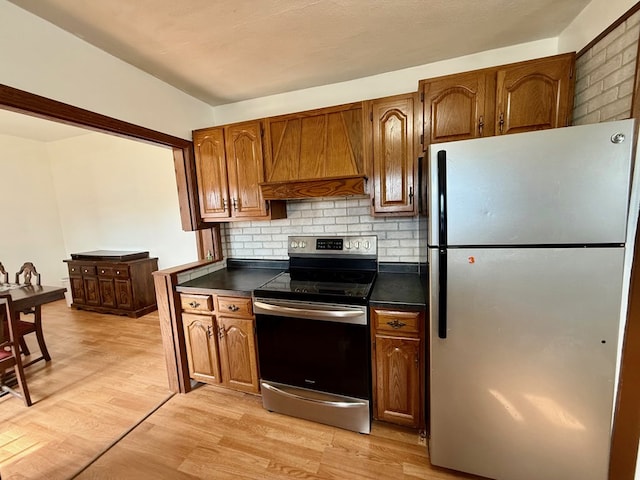 kitchen with brown cabinets, light wood finished floors, dark countertops, appliances with stainless steel finishes, and premium range hood