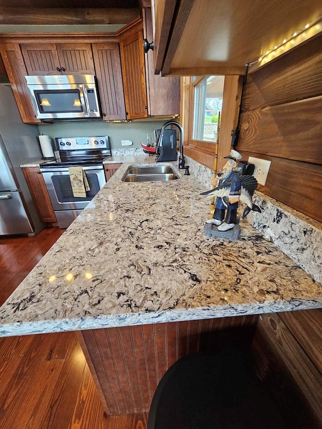 kitchen with stainless steel appliances, a sink, dark wood-style flooring, brown cabinets, and light stone countertops