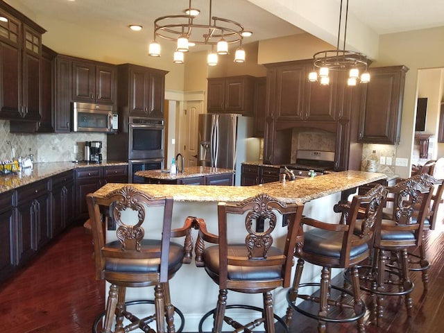 kitchen with decorative light fixtures, stainless steel appliances, light stone countertops, dark brown cabinets, and a center island with sink