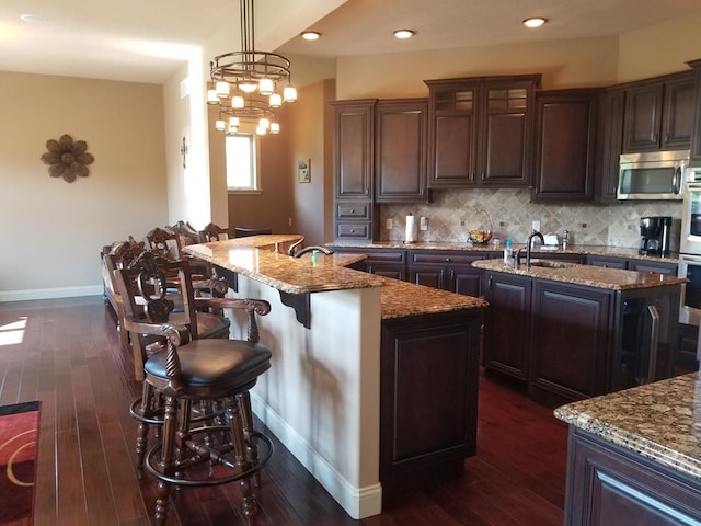 kitchen with pendant lighting, dark brown cabinets, stainless steel appliances, light stone countertops, and an island with sink