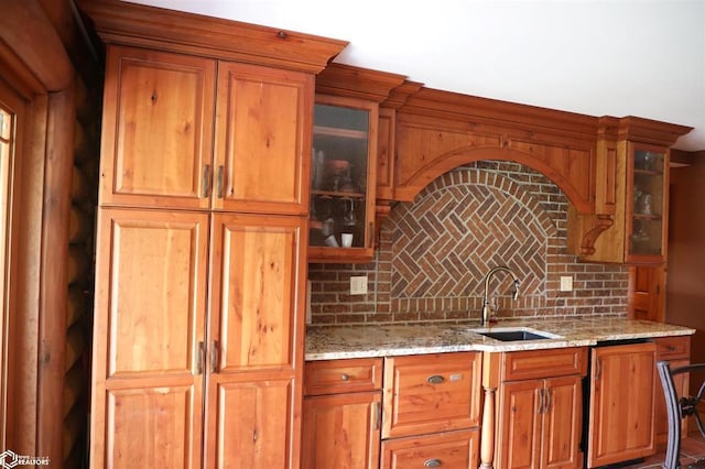 kitchen featuring light stone counters, a sink, backsplash, brown cabinets, and glass insert cabinets