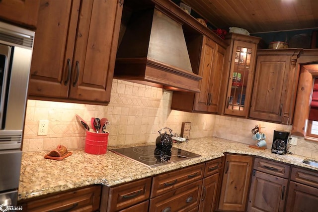 kitchen featuring light stone counters, tasteful backsplash, custom range hood, glass insert cabinets, and black electric cooktop