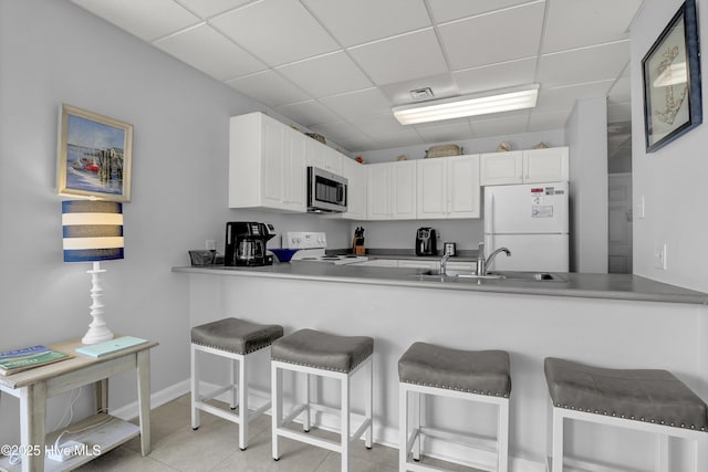 kitchen with a sink, white cabinets, a peninsula, a drop ceiling, and white appliances