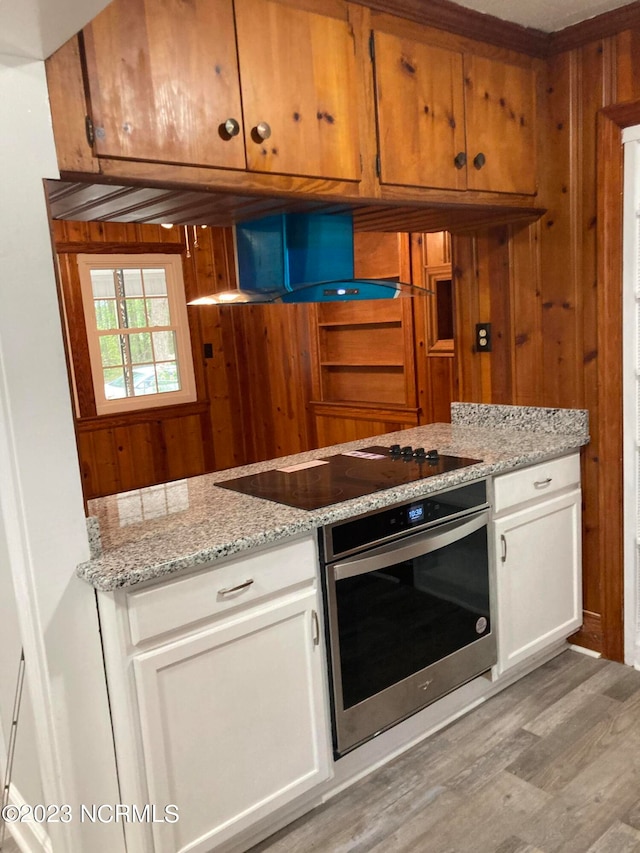 kitchen with white cabinets, wall chimney exhaust hood, black electric cooktop, light wood-type flooring, and stainless steel oven