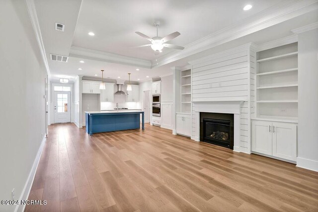 unfurnished living room featuring crown molding, light wood-type flooring, a raised ceiling, built in features, and ceiling fan