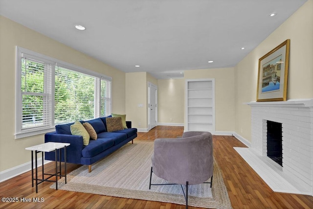 living room featuring built in shelves, wood finished floors, a brick fireplace, baseboards, and recessed lighting