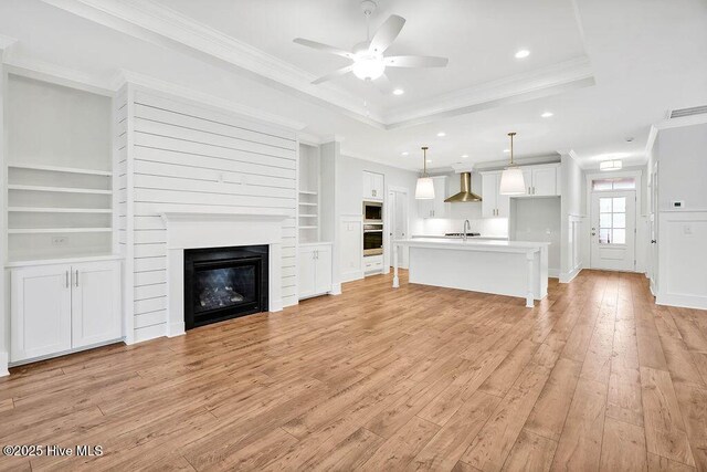 unfurnished living room with light hardwood / wood-style flooring, built in features, ceiling fan, ornamental molding, and a raised ceiling