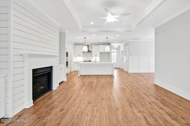 unfurnished living room featuring crown molding, ceiling fan, a raised ceiling, and light wood-type flooring