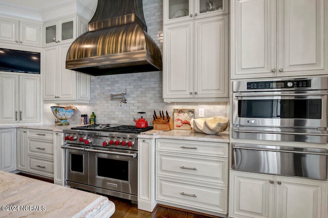 kitchen with light stone counters, stainless steel appliances, custom exhaust hood, tasteful backsplash, and white cabinetry