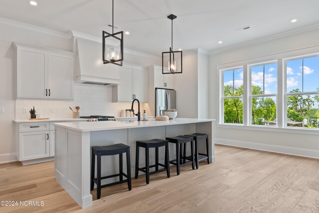 kitchen featuring decorative light fixtures, custom exhaust hood, an island with sink, stainless steel fridge, and light wood-type flooring