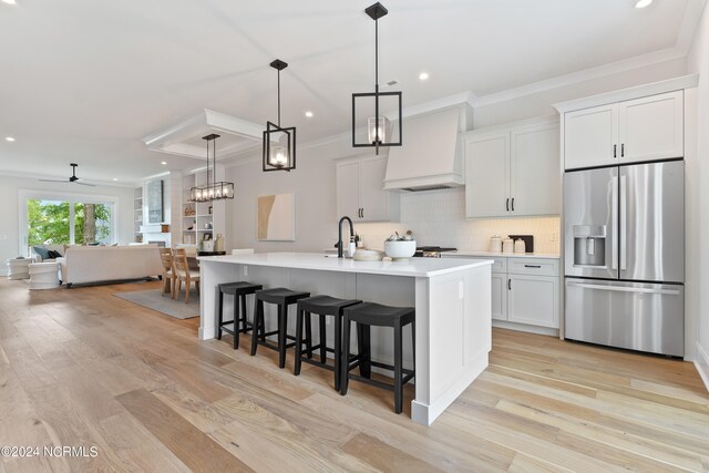 kitchen with light hardwood / wood-style floors, custom range hood, an island with sink, decorative light fixtures, and stainless steel refrigerator with ice dispenser