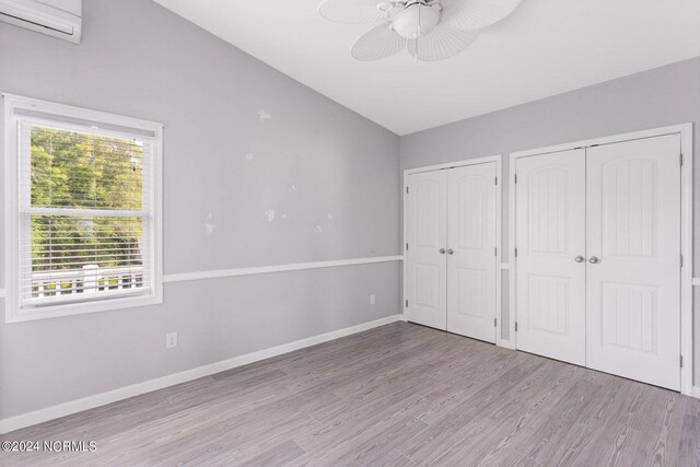 unfurnished bedroom with vaulted ceiling, a wall mounted AC, ceiling fan, light hardwood / wood-style floors, and multiple closets