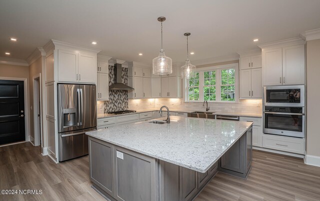 kitchen with wall chimney range hood, backsplash, an island with sink, wood-type flooring, and appliances with stainless steel finishes