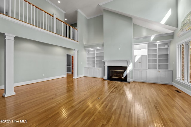 unfurnished living room featuring decorative columns, light wood-type flooring, built in shelves, a high ceiling, and crown molding