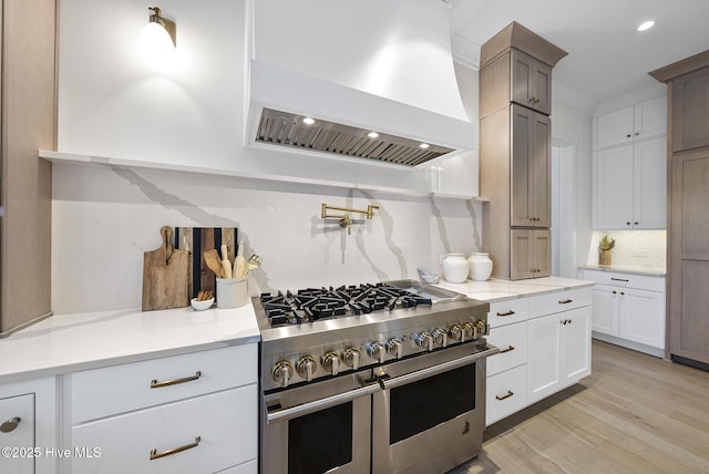 kitchen with custom range hood, double oven range, backsplash, white cabinetry, and light wood finished floors