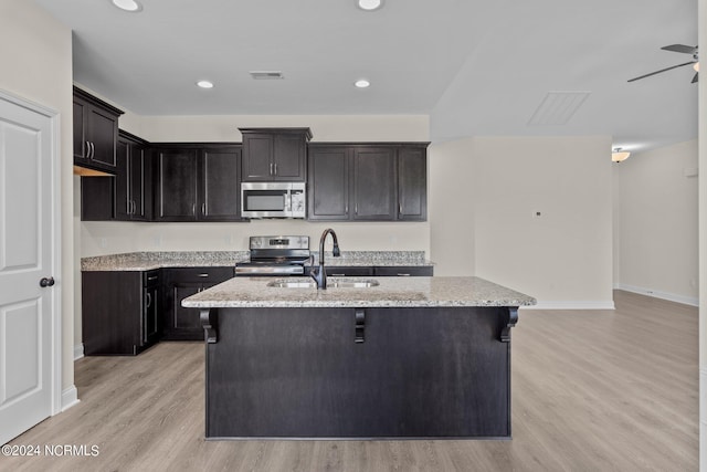 kitchen with a center island with sink, sink, light wood-type flooring, light stone countertops, and appliances with stainless steel finishes