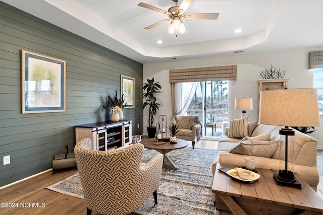 living room with ceiling fan, wood walls, wood-type flooring, and a tray ceiling