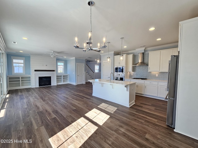 kitchen with an island with sink, hanging light fixtures, stainless steel appliances, crown molding, and wall chimney exhaust hood