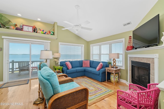 living room featuring light hardwood / wood-style flooring, vaulted ceiling, a water view, a tiled fireplace, and ceiling fan