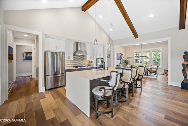 kitchen featuring white cabinets, decorative light fixtures, wall chimney range hood, an island with sink, and stainless steel fridge