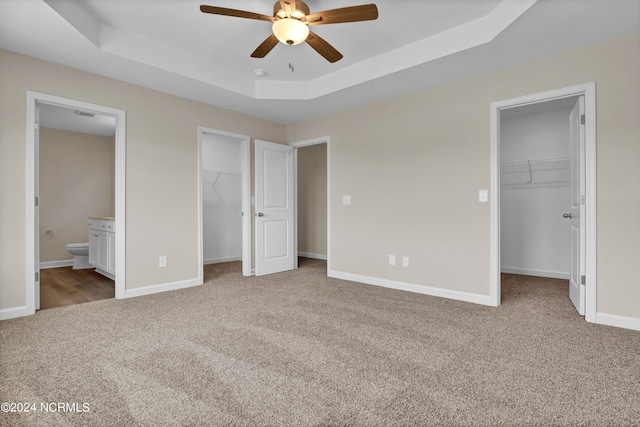 unfurnished bedroom with a spacious closet, a tray ceiling, ceiling fan, connected bathroom, and light colored carpet