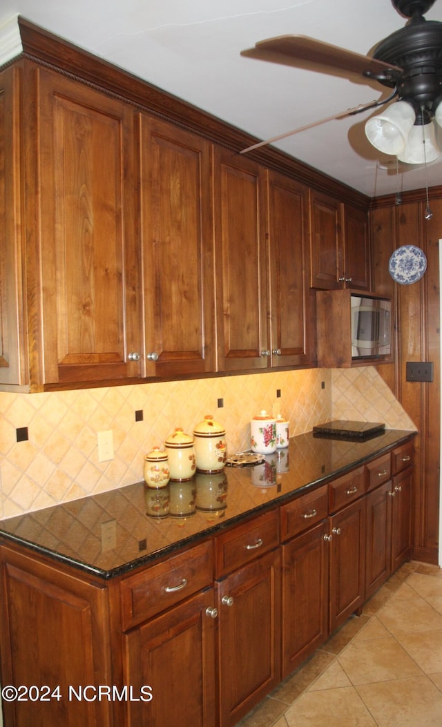 kitchen featuring light tile flooring, backsplash, stainless steel microwave, dark stone countertops, and ceiling fan