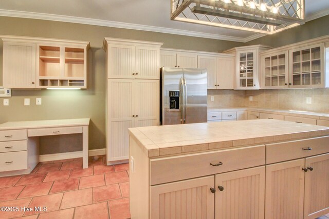 kitchen with ornamental molding, stainless steel refrigerator with ice dispenser, open shelves, built in desk, and glass insert cabinets