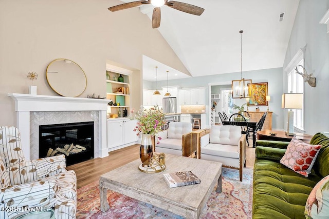 living room featuring ceiling fan with notable chandelier, high vaulted ceiling, a tiled fireplace, built in shelves, and light wood-type flooring