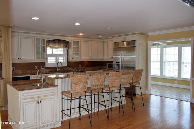 kitchen featuring built in refrigerator, dark stone countertops, ornamental molding, an island with sink, and light hardwood / wood-style floors