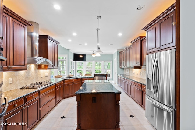 kitchen with wall chimney range hood, dark stone counters, stainless steel appliances, ceiling fan, and a center island