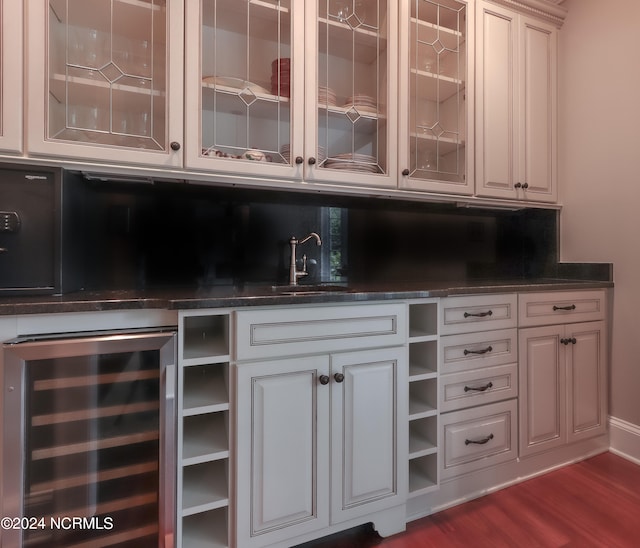 bar with beverage cooler, dark stone countertops, dark wood-type flooring, sink, and white cabinets