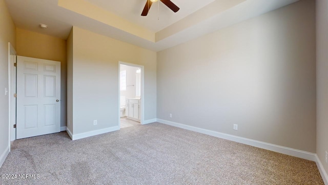 unfurnished bedroom featuring a tray ceiling, ensuite bath, ceiling fan, and light colored carpet