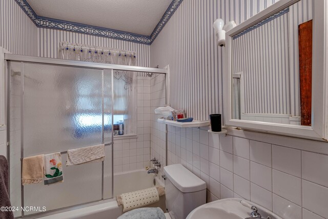 bathroom featuring a textured ceiling, toilet, bath / shower combo with glass door, tile walls, and wallpapered walls