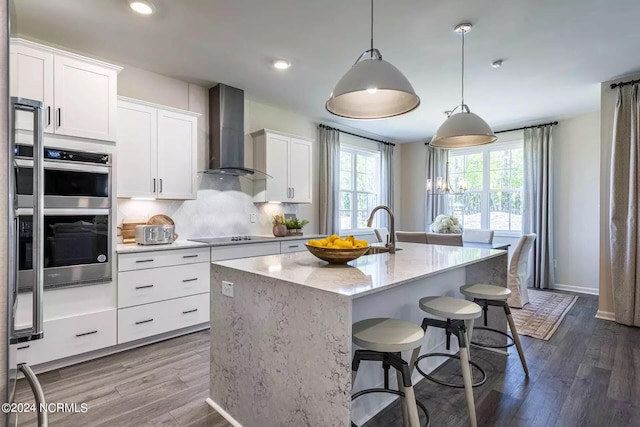 kitchen with wall chimney range hood, a kitchen island with sink, double oven, hardwood / wood-style floors, and backsplash