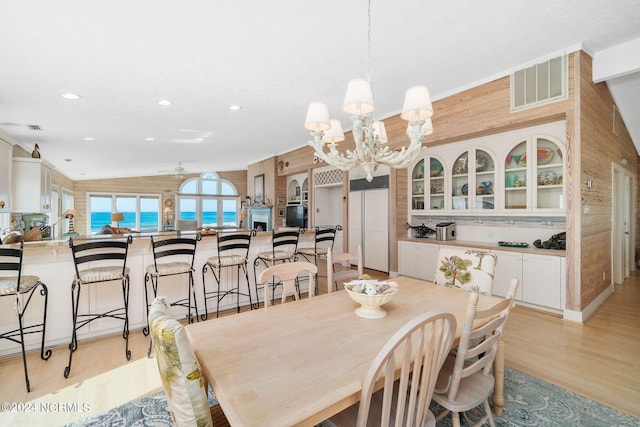 dining room with vaulted ceiling, wooden walls, light hardwood / wood-style flooring, and ceiling fan with notable chandelier