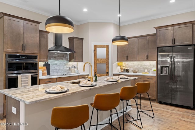 kitchen featuring pendant lighting, stainless steel appliances, a center island with sink, and wall chimney range hood