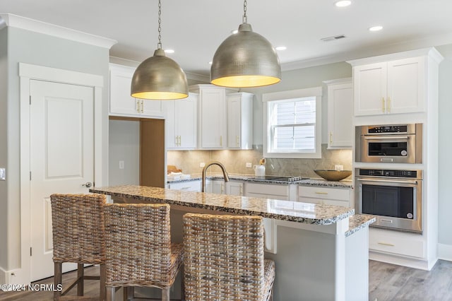 kitchen featuring pendant lighting, a kitchen island with sink, light stone countertops, white cabinets, and decorative backsplash