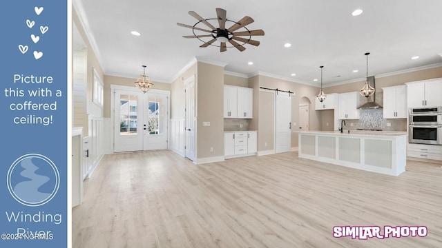 kitchen with double oven, wall chimney range hood, a barn door, a center island with sink, and white cabinets